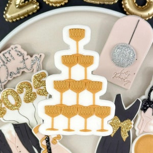 Könnte beinhalten: Ein schwarzer Teller mit goldenen und schwarzen Keksen zum Thema Silvester. Die Kekse beinhalten einen Champagnerglas-Turm, einen Smoking, ein Kleid, eine Champagnerflasche, einen Ballon und ein Schild mit der Aufschrift "Let's Party".