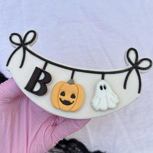 Könnte beinhalten: Ein weißer Keks, der mit schwarzem Zuckerguss in Form eines Banners mit dem Wort "BOO" und Halloween-Dekorationen verziert ist, darunter ein Kürbis und ein Geist.