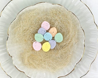 May include: A white plate with a gold rim and a nest of shredded straw inside. The nest contains pastel-colored candy eggs that spell out "Frohe Ostern" in German.