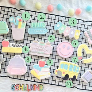 Könnte beinhalten: Eine Sammlung farbenfroher Kekse zum Thema Schule. Die Kekse umfassen einen Bleistift und eine Schere, ein Schild mit der Aufschrift "Hallo 1. Klasse", ein Banner mit Buntstiften, ein Lineal mit einem Apfel, ein Smiley, einen herzförmigen Bleistift, einen Rucksack, Bücher mit einem Apfel und einen Schulbus.