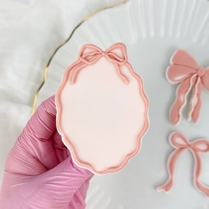 Può includere: Tre decorazioni per biscotti in pasta di zucchero rosa. Una è una cornice rotonda con un fiocco rosa in cima. Le altre due sono fiocchi rosa con code.