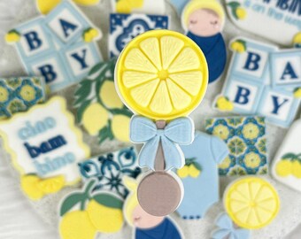 May include: A collection of blue and yellow decorated sugar cookies with various baby-themed designs, including a rattle, a baby bottle, a baby onesie, and blocks that spell "BABY". The cookies are decorated with lemon slices and the text "Il mio bambino e' arrivato" is on one of the cookies.