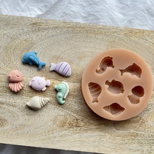 Puede incluir: Un molde de silicona con varias formas de criaturas marinas, incluyendo un delfín, un pez, una concha y una estrella de mar. También hay varios amuletos de criaturas marinas de resina de colores junto al molde.