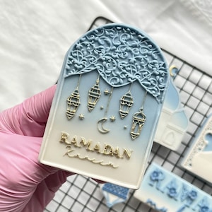 Peut inclure: Un biscuit bleu et blanc décoré de détails dorés. Le biscuit est en forme d'arche et présente un croissant de lune, des étoiles et des lanternes. Le texte "RAMADAN Kareem" est écrit en lettres dorées.