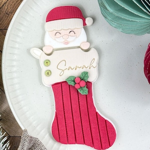 Puede incluir: Una galleta de Santa Claus roja y blanca en una galleta de calcetín roja con el nombre "Sarah" escrito en ella.