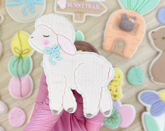 May include: A collection of Easter-themed sugar cookies decorated with pastel colors and various designs, including a lamb, a bunny, a basket of eggs, and a flower. The cookies are arranged on a wooden surface.