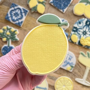 Könnte beinhalten: Ein gelber Zitronenkeks mit einem grünen Blatt oben drauf. Der Keks ist umgeben von anderen Keksen, darunter ein Zitronenbaum, ein Glas Limonade und Fliesen mit blauen und weißen Mustern.