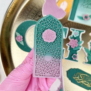 Könnte beinhalten: Ein rosa-grüner Dekorationskeks mit einem komplizierten geometrischen Muster und arabischer Kalligraphie. Der Keks ist in Form eines Fensters oder eines Bogens geformt.
