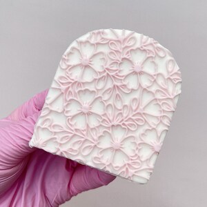 Peut inclure: Un outil de décoration de gâteau en silicone blanc avec un motif floral rose. L'outil est en forme d'arche.