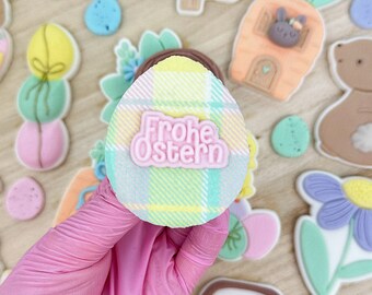 May include: A hand holding a pastel yellow, pink, and green plaid Easter egg cookie with the German phrase "Frohe Ostern" in pink lettering. The cookie is surrounded by other Easter-themed cookies, including a bunny, a carrot, and a basket of eggs.