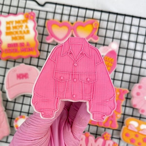 May include: A collection of pink iced cookies, including a jacket, sunglasses, a baseball cap, and a sign that reads "My mom is not a regular mom, she is a cool mom." The cookies are arranged on a wire rack.