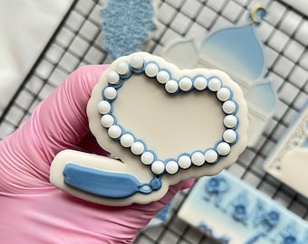 May include: A blue and white cookie decorated with a heart-shaped string of white beads. The cookie is on a pink gloved hand. The cookie is part of a set of cookies with a Ramadan theme.