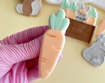 May include: A hand holding a light orange carrot cookie with green icing on top. The cookie is decorated with a light brown stem and green leaves. The cookie is on a white surface.