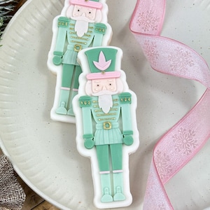 May include: Two green and white gingerbread cookies decorated with gold details and pink accents, shaped like nutcracker soldiers. The cookies are on a white plate with a pink ribbon.