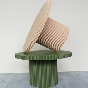 May include: A modern, abstract composition featuring three cylindrical tables. The bottom table is olive green, supporting a light pink table with a ridged edge. A third table, also light pink, rests on the second table. The tables have a minimalist design.