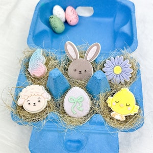 Op de afbeelding: Een blauwe eierdoos gevuld met pastelkleurige paas-koekjesdecoraties. De decoraties omvatten een konijn, een lam, een kuiken, een vlinder, een ei en een bloem.