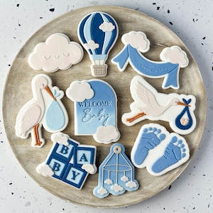 Può includere: Una collezione di biscotti a tema baby disposti su un vassoio di legno. I biscotti includono una mongolfiera, nuvole, una cicogna con un bambino, blocchi per bambini e impronte. I biscotti sono decorati in tonalità di blu e bianco.