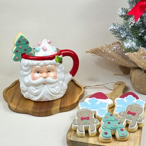 Può includere: Immagine a tema natalizio con una tazza di Babbo Natale piena di panna montata e un biscotto a forma di albero. Accanto, un tagliere in legno con biscotti a forma di omino di pan di zenzero e fiocco di neve. Un piccolo albero di Natale con un fiocco rosso sullo sfondo.