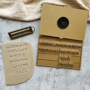Può includere: Un set di taglia biscotti color oro con un manico rettangolare, una custodia e un biscotto campione. La custodia contiene timbri di lettere individuali. Il biscotto campione mostra lettere maiuscole e minuscole, numeri e simboli. Il testo "COOKIECUTTERFABRIK" è visibile.