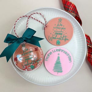 Könnte beinhalten: Weihnachtliche Dekoration mit einer transparenten Kugel mit grüner Schleife, gefüllt mit Konfetti und Text. Zwei runde Ornamente mit deutschem Text und Weihnachtsbaum-Designs befinden sich ebenfalls auf einem weißen Teller.