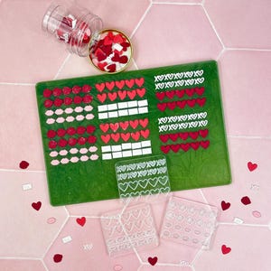 Könnte beinhalten: Ein Bild zum Valentinstag mit einem grünen Tablett mit roten Herzen, Lippen und XOXO-Designs. Klare Formen mit Herz- und Lippenformen sind ebenfalls vorhanden, zusammen mit verstreuten herzförmigen Süßigkeiten und einem Glas voller Süßigkeiten. Der Hintergrund ist eine rosa sechseckige Fliese.