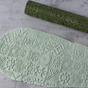 Könnte beinhalten: Ein olivgrüner, gemusterter Nudelholz und ein Stück hellgrüner Ton mit einem geprägten Design. Der Ton zeigt ein detailliertes, sich wiederholendes Muster aus geometrischen und floralen Formen, das durch das Nudelholz erzeugt wurde.