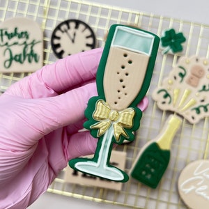 Può includere: Un biscotto di zucchero verde e oro a forma di flauto di champagne. Il biscotto è decorato con un fiocco dorato e ha una farcitura marrone chiaro. Il biscotto è su una griglia.