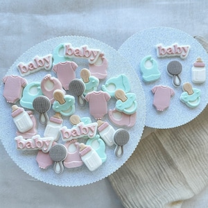 May include: Two white plates with blue glitter, each decorated with pink and blue fondant baby shower decorations. The decorations include baby bottles, pacifiers, onesies, and the word "baby".