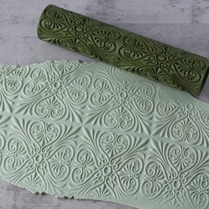 Op de afbeelding: Een olijfgroene deegroller met een gedetailleerd bloemmotief ligt naast een kleiblad met hetzelfde reliëf. De klei heeft een lichtgroene kleur en toont de afdruk van het ontwerp van de deegroller. De deegroller is cilindrisch.