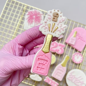 Könnte beinhalten: Ein rosa und goldener Zuckerkeks, verziert mit einer Champagnerflasche, einem herausspringenden Korken und den Worten "Happy New Year".