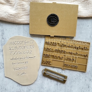 Könnte beinhalten: Ein goldfarbenes Ausstechersets mit Groß- und Kleinbuchstaben, Zahlen und Symbolen. Das Set enthält einen Stempel der Buchstaben und Zahlen auf einer beigen Oberfläche. Der Text "COOKIECUTTERFABRIK" ist sichtbar auf dem Ausstecher.