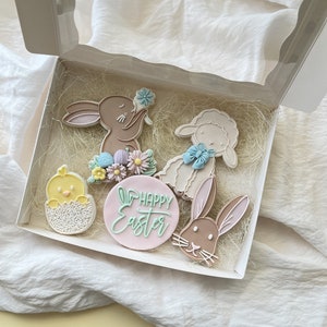 Op de afbeelding: Een geschenkdoos gevuld met paas-suikerkoekjes. De koekjes zijn versierd met pastelkleuren en bevatten een kuiken, konijn, lam en een roze koekje met de woorden "Happy Easter" in groene letters.