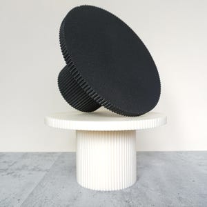 Puede incluir: Una escultura abstracta moderna con un gran disco negro texturizado y un cilindro negro más pequeño y acanalado. Descansa sobre un pedestal cilíndrico blanco con ranuras verticales, creando un contraste llamativo.