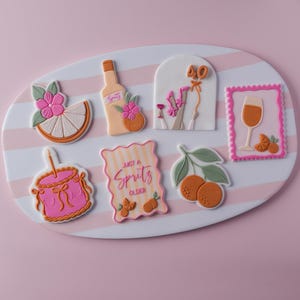 May include: A set of seven decorated biscuits featuring an orange slice with a pink flower, a spritz bottle, an arch with the number 40, a wine glass in a pink frame, a pink cake with a candle, and a sign reading Just a Spritz Older with oranges.