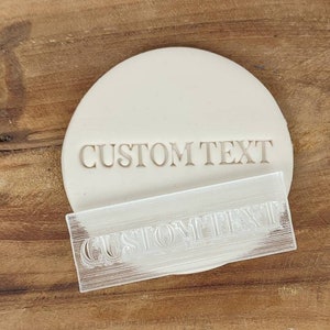 Può includere: Un timbro per biscotti in plastica trasparente con le parole "CUSTOM TEXT" incise su di esso. Il timbro è su una superficie di legno con le parole "Cookie Cutter Fab" incise su di essa.