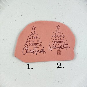 Könnte beinhalten: Hellrosa, baumförmige Weihnachtsdekoration mit dem Text "We wish you a Merry Christmas" und "Wir wünschen euch Frohe Weihnachten" in Schwarz. Die Dekoration befindet sich auf einem weißen Teller.