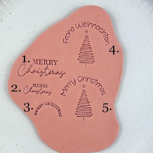 Könnte beinhalten: Ein weißer Keramikteller mit strukturiertem Rand hält ein korallenfarbenes, unregelmäßig geformtes Stück. Das Korallenstück zeigt die Wörter "Merry Christmas" und "Frohe Weihnachten" in einer Schriftart, zusammen mit Weihnachtsbaum-Illustrationen.