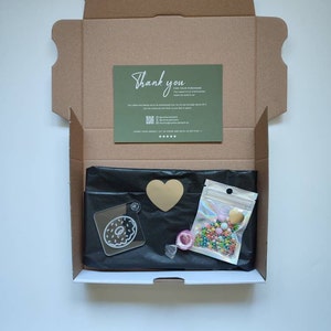 May include: A brown cardboard box with a thank you card inside. The box contains a black bag with a donut-shaped acrylic charm, a gold heart, a small bag of colorful sprinkles, and a roll of clear tape.