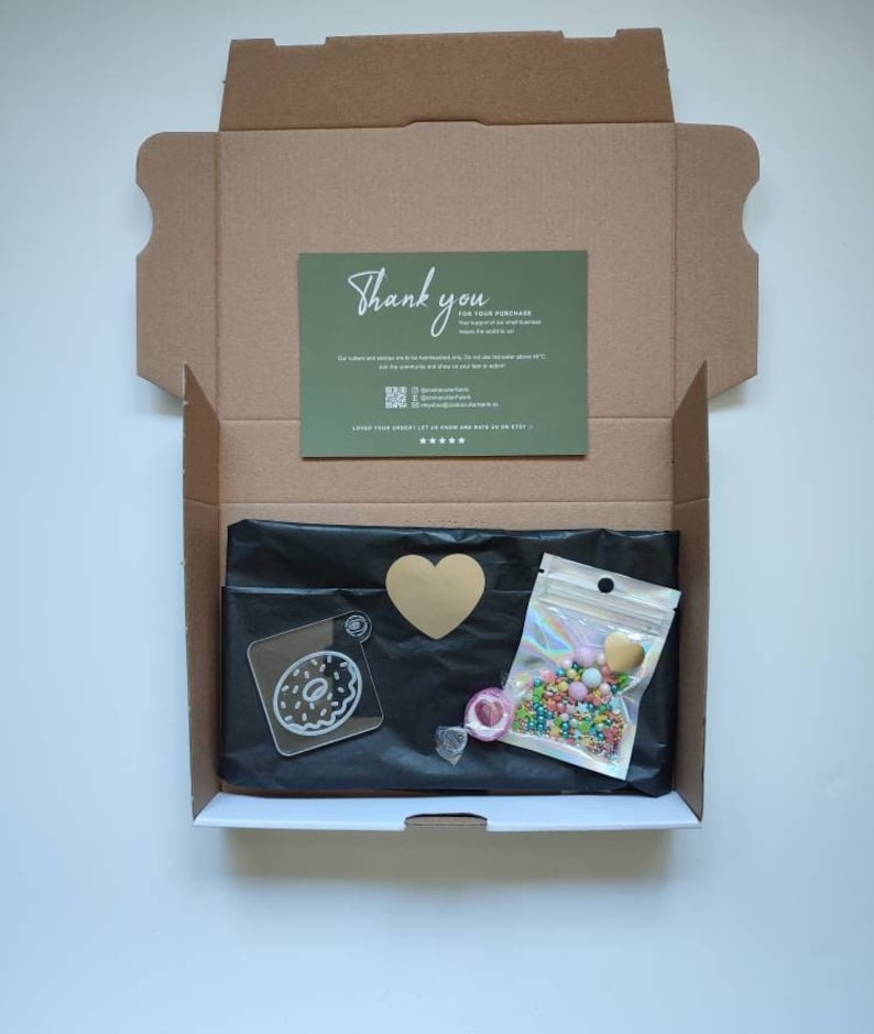 May include: A brown cardboard box with a thank you card inside. The box contains a black bag with a donut-shaped acrylic charm, a gold heart, a small bag of colorful sprinkles, and a roll of clear tape.