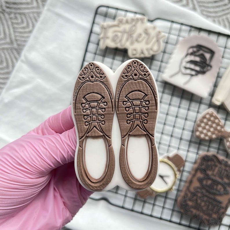 May include: Two brown rubber stamps shaped like shoes with a detailed design. The stamps are held in a hand wearing a pink glove.