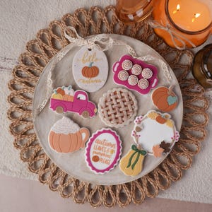 Könnte beinhalten: Eine Sammlung dekorativer Kekse mit Herbstmotiven, darunter ein rosa Lastwagen mit Kürbissen, ein Gitterkuchen, eine kürbisförmige Tasse, ein Schild mit der Aufschrift hello autumn, ein Tablett mit Zimtschnecken, ein Kürbis und eine Plakette.
