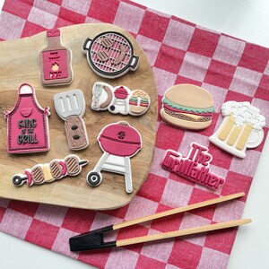 May include: A set of nine colourful, hand-painted cookie cutters shaped like grilling and barbecue-themed items, including a grill, a burger, a beer mug, a hot dog, and a grill master's hat. The cookie cutters are arranged on a wooden chopping board with a red and white checked tablecloth.