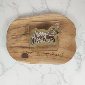 May include: A gold plastic cookie cutter in the shape of the words "Merry Christmas" with a small Santa hat on top. The cookie cutter is on a wooden cutting board.