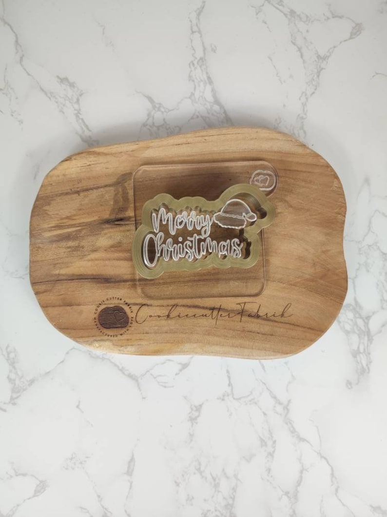 May include: A gold plastic cookie cutter in the shape of the words "Merry Christmas" with a small Santa hat on top. The cookie cutter is on a wooden cutting board.
