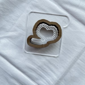 May include: A clear plastic sheet with a brown wooden cutout of a heart shape with a white pearl necklace inside.