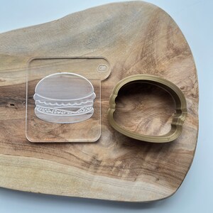 May include: A clear acrylic cookie cutter with a burger design and a gold metal cookie cutter in the shape of a burger bun.