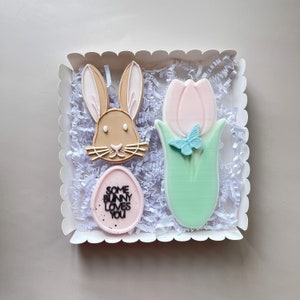 May include: A white gift box filled with pastel-coloured Easter cookies. The cookies are shaped like a bunny, a tulip, and an egg. The egg cookie has the text "Some Bunny Loves You" on it.