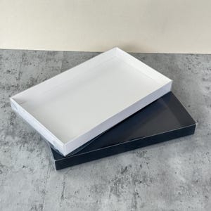 May include: Two rectangular gift boxes, one white and one black, are stacked. The white box is on top, angled to reveal the black box beneath. Both boxes have a glossy finish and are empty. Ideal for packaging.