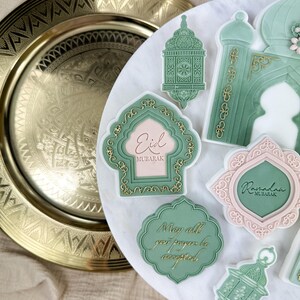 May include: A set of green and pink decorated cookies with gold accents. The cookies feature Islamic designs and text, including "Eid Mubarak" and "Ramadan Mubarak". One cookie reads "May all your prayers be accepted".