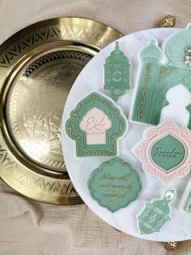 May include: A set of green and pink decorated cookies with gold accents. The cookies feature Islamic designs and text, including "Eid Mubarak" and "Ramadan Mubarak". One cookie reads "May all your prayers be accepted".